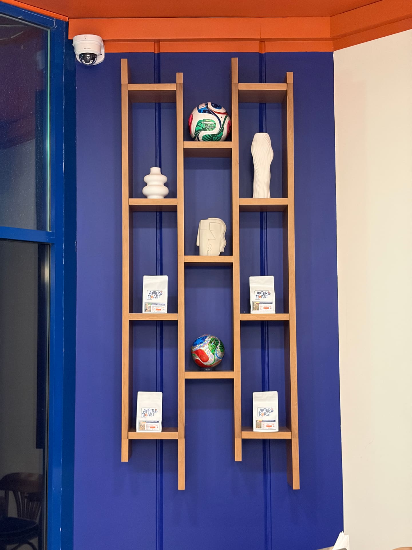 Wooden ladder shelving on a cobalt wall holding four white After Roast retail coffee bags, each with a country label, alongside footballs and ceramics.
