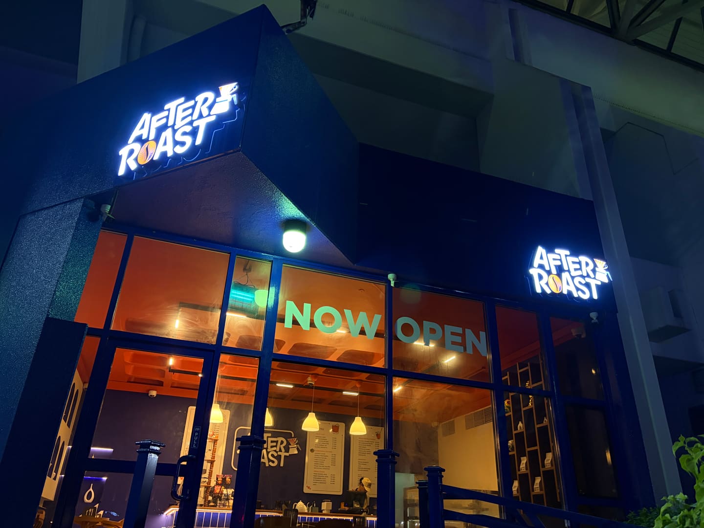 The After Roast storefront at night, two illuminated signs above tall blue-framed windows lettered NOW OPEN, the lit room visible inside.